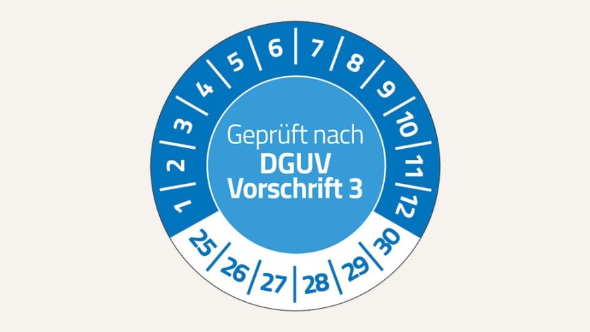 Man sieht eine runde Prüfplakette. In der Mitte steht der Text "Geprüft nach DGUV Vorschrift 3". Im oberen Bereich stehen im Halbkreis die Zahlen 1 bis 12. Unten stehen die Zahlen 25 bis 30.
