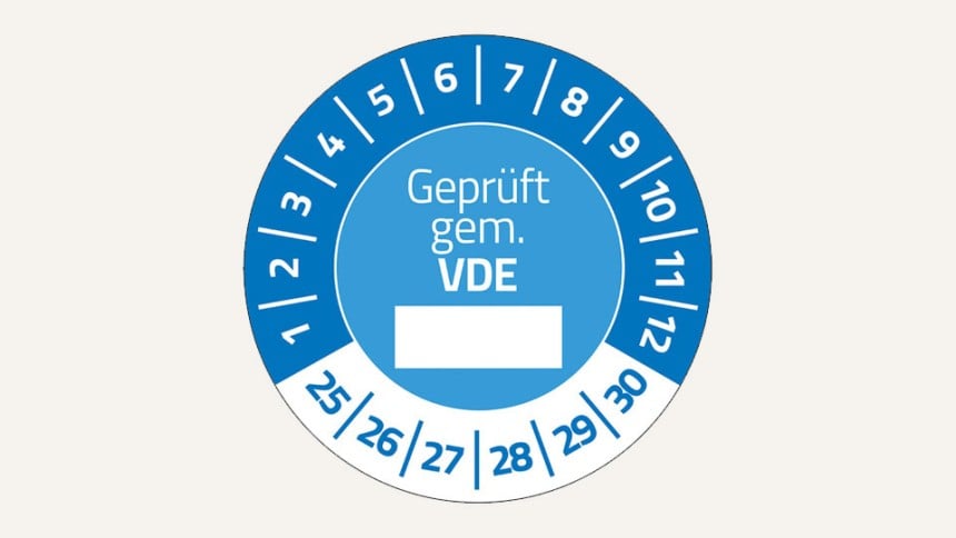 Man sieht eine blaue Prüfplakette. In der Mitte steht "Geprüft gem. VDE", darunter ist ein weißer Kasten, in den man etwas eintragen kann. Im oberen Bereich stehen im Halbkreis die Zahlen 1 bis 12. Unten im Halbkreis die Zahlen 25 bis 30.