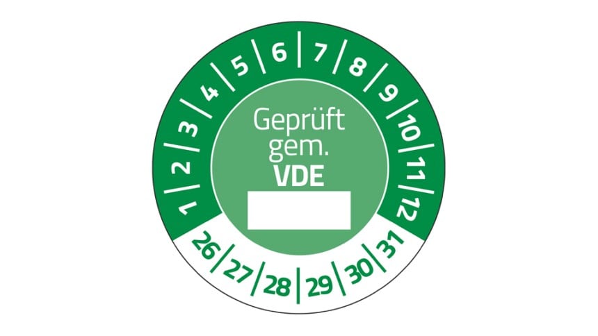 Eine grüne Prüfplakette. In der Mitte steht "Geprüft gem. VDE", darunter ein weißer Kasten. Im Halbkreis stehen die Zahlen 1 bis 12, unten die Zahlen 26 bis 31.
