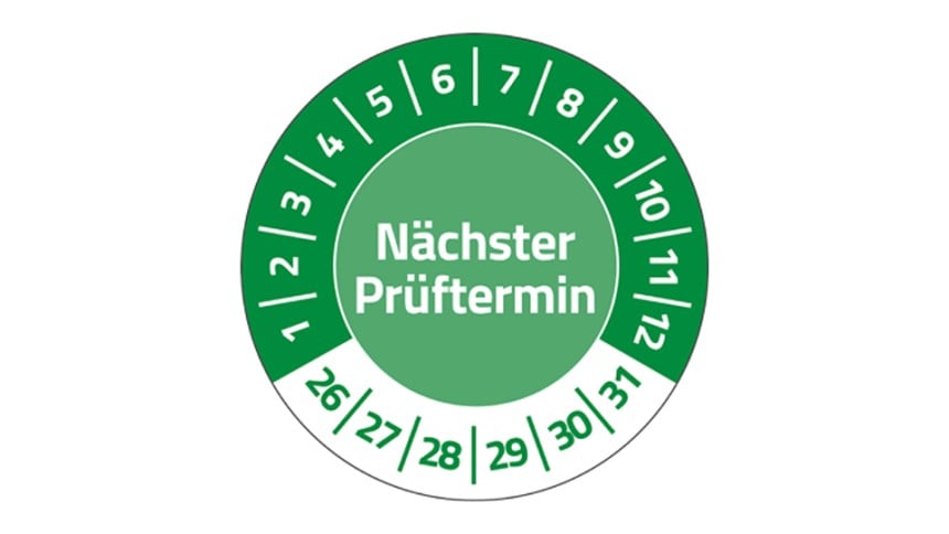 Eine grüne Prüfplakette, in deren Mitte steht "Nächster Prüftermin". Im Halbkreis stehen die Zahlen 1 bis 12. Unten stehen die Zahlen 26 bis 31.