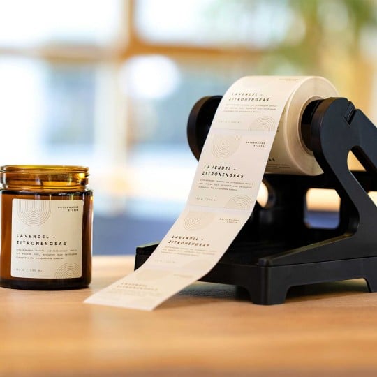 Neben einem Rollenetiketten-Abroller, auf dem eine Rolle mit bedruckten Etiketten aufgerollt ist, steht eine Kerze, auf der eines dieser Etiketten klebt.