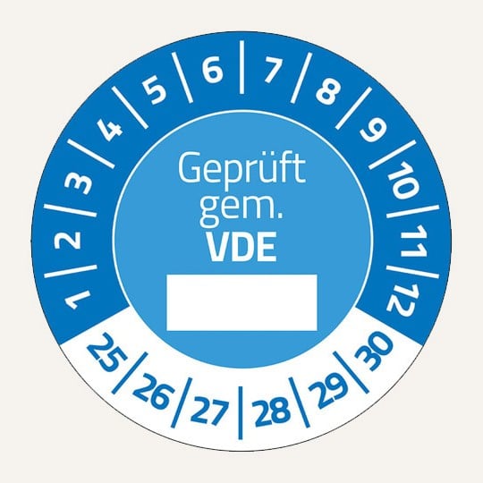 Man sieht eine blaue Prüfplakette. In der Mitte steht "Geprüft gem. VDE", darunter ist ein weißer Kasten, in den man etwas eintragen kann. Im oberen Bereich stehen im Halbkreis die Zahlen 1 bis 12. Unten im Halbkreis die Zahlen 25 bis 30.