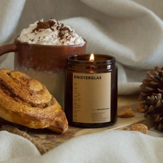 Eine Kerze mit bedrucktem Aufkleber steht neben einem Kaffee und einer Nussschnecke. Auf dem Aufkleber steht "Knisterglas".