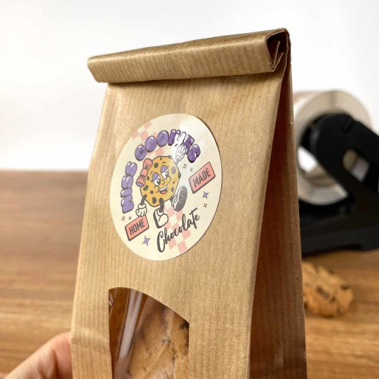 Auf einer braunen Papiertüte, in der Kekse sind, klebt ein runder Aufkleber. Auf diesem steht "Miss Cookies", das mit Spotlack veredelt ist.