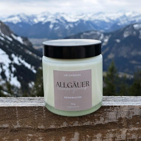 Das Bild zeigt im Vordergrund ein Glas Kerzenwachs samt Etikett und im Hintergrund sind beschneite Berge zu erkennen.