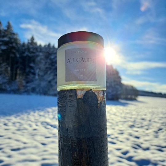 Das Bild zeigt im Vordergrund ein Glas mit Kerzenwachs samt Etikett und im Hintergrund eine verschneite Wiese und Wald.