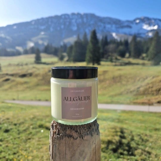 Das Bild zeigt ein Glas Kerzenwachs samt Etikett das im Vordergrund auf einem Holzstamm steht. Im Hintergrund ist eine Wiese und ein Berg erkennbar.