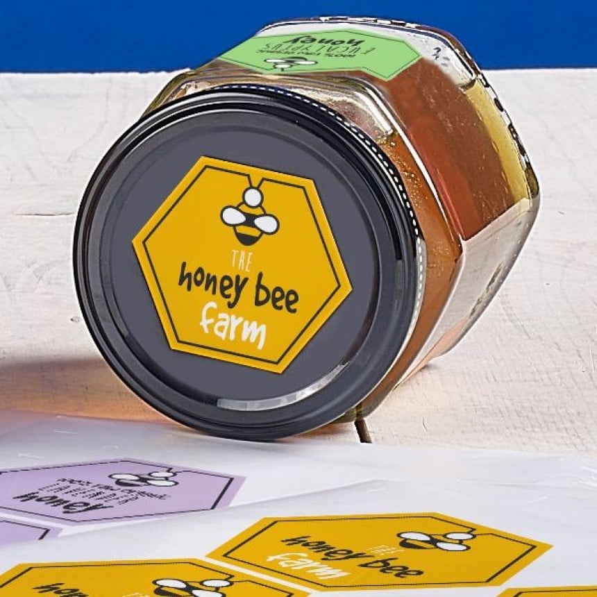 Auf dem Deckel eines Honigglases klebt ein sechseckiges, bedrucktes Honigetikett. Darauf steht "The honey bee farm".