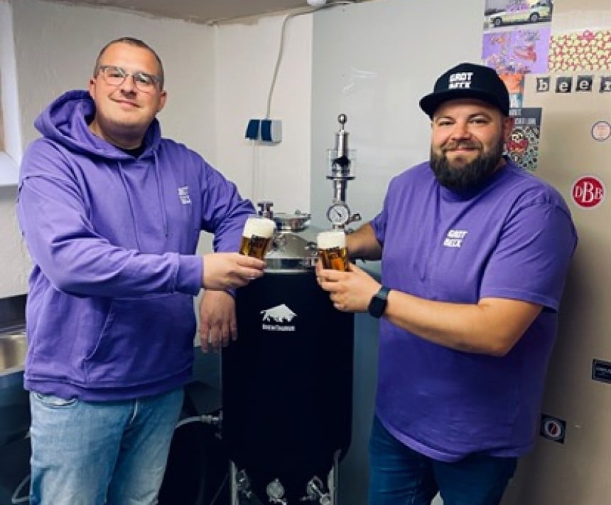 Zwei Menschen stehen mit einem Bierglas vor ihrer kleinen Brauereimaschine.