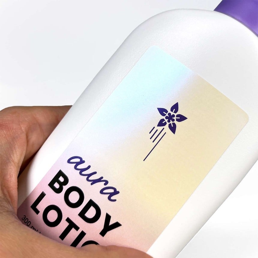 Auf einem Body Lotion Behältnis klebt ein bedruckter Aufkleber aus Perlmutt-Folie. Auf dem Aufkleber steht "aura Body Lotion" und eine Blume ist abgebildet. Der Fokus des Bildes liegt auf dem Material des Aufklebers.