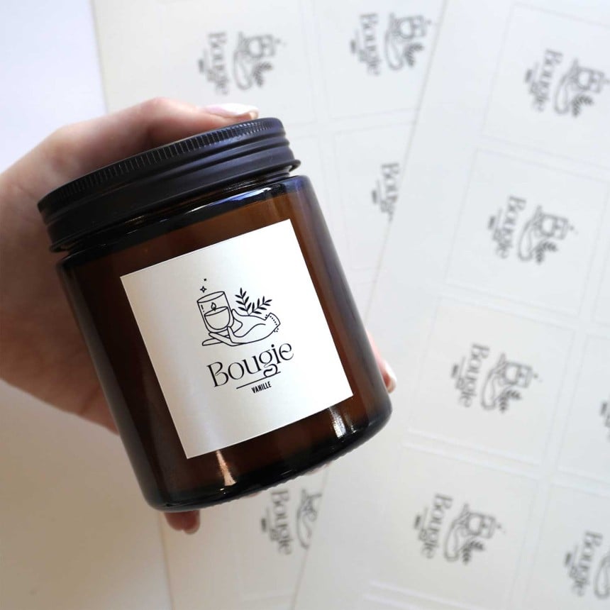 Im Vordergrund hält eine Hand ein Glasbehältnis auf dem ein bedruckter Aufkleber aus Perlmutt Folie klebt. Im Hintergrund sieht man die Aufkleber auf verschiedenen Bögen.