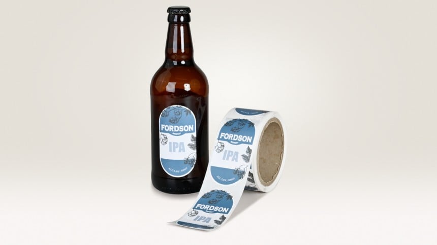 Neben einer Bierflasche, auf der ein blau/weißes Etikett klebt, liegt eine Rolle, auf der ebendiese bedruckten Rollenetiketten kleben.