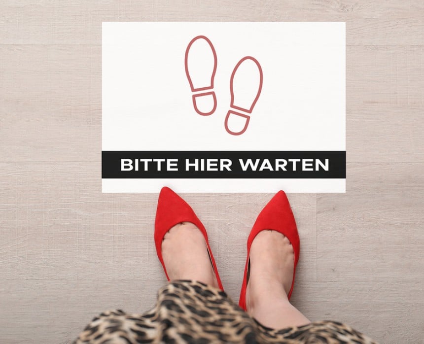 Auf dem Boden klebt ein Bodenaufkleber auf dem zwei Schuhabdrücke zu sehen sind und "Bitte hier warten" steht. Davor steht eine Frau.