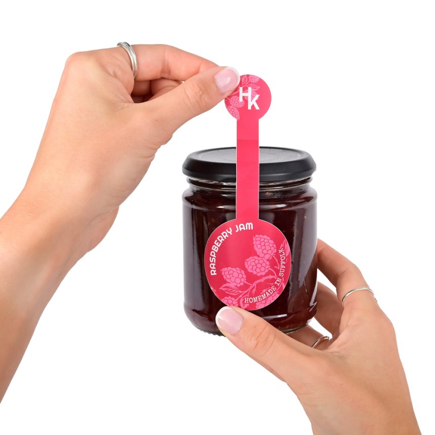 Jemand klebt ein bedrucktes Verschlussetikett mit zwei runden Enden auf ein Marmeladenglas.