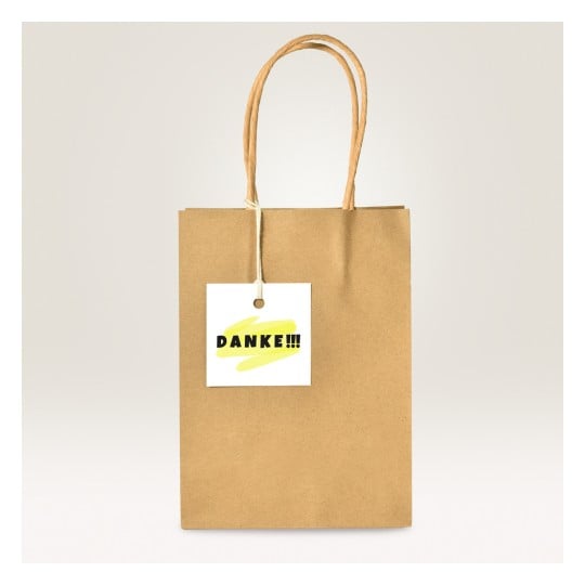Das Bild zeigt eine braune Papiertüte mit einem Etikett, auf dem 'DANKE!!!' steht, das ein gelbes Pinselstrichdesign aufweist.