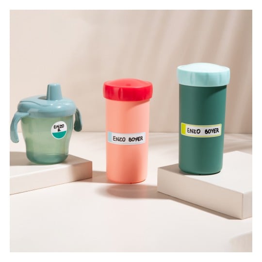 Three colorful beverage containers arranged on a surface. The left container is a sippy cup, the middle is a peach-colored drink container, and the right is a dark green tumbler, all labeled with 'ENZO BOYER'.