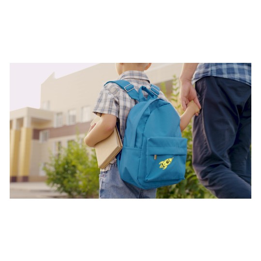 Ein junges Kind mit einem blauen Rucksack mit Raketenmotiv hält ein Buch in der einen Hand und die Hand eines Erwachsenen in der anderen, während es auf einem Weg in der Nähe von Gebäuden und Grünflächen geht.