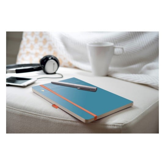 Das Bild zeigt ein blaues Notizbuch mit einem orangefarbenen Band, einen darauf liegenden Stift, in der Nähe gelegene Kopfhörer, ein Smartphone und eine weiße Tasse, alles auf einer hellen Couch mit einer strukturierten Decke im Hintergrund.