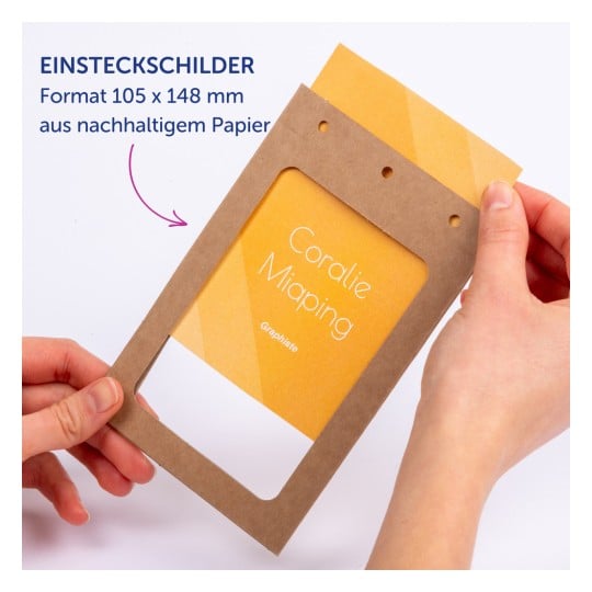 Das Bild zeigt eine Person, die einen Rahmen aus braunem Papier mit einer gelben Karte darin hält. Der sichtbare Text umfasst 'Coralie Miaping' und Informationen über das Material, die darauf hinweisen, dass es sich um nachhaltiges Papier handelt. Auch die Abmessungen sind angegeben.