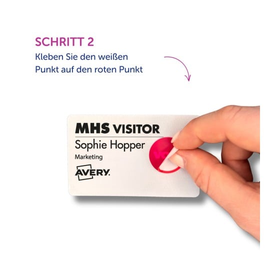 Das Bild zeigt eine Hand, die einen weißen Aufkleber auf einem Besucher-Ausweis anbringt, der den Namen Sophie Hopper und den Bereich Marketing enthält. Es enthält Anweisungen auf Deutsch, einen weißen Punkt auf einen roten Punkt zu setzen.