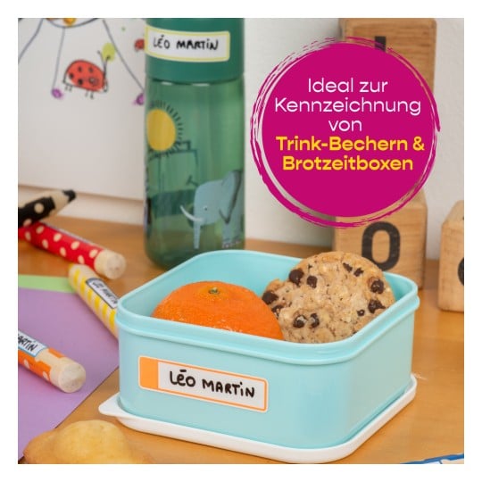 Das Bild zeigt eine blaue Lunchbox, die eine Orange und Kekse enthält, neben einer Wasserflasche, Buntstiften und Holzklötzen.