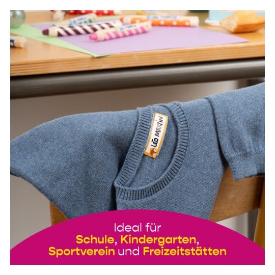 Das Bild zeigt einen blauen Pullover, der auf einem Stuhl hängt, mit bunten Markern und Papieren, die im Hintergrund auf dem Tisch sichtbar sind. Ein Etikett auf dem Pullover weist darauf hin, dass er ideal für Schule, Kindergarten und sportliche Aktivitäten ist.