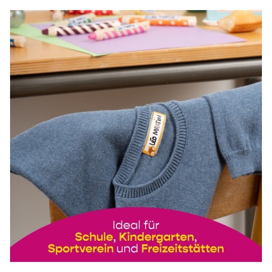Ein blauer Pullover mit einem Etikett auf einem Stuhl in der Nähe eines Tisches mit bunten Wachsmalstiften und Papierbögen. Das Bild deutet auf Eignung für Schul- und Freizeitaktivitäten hin.