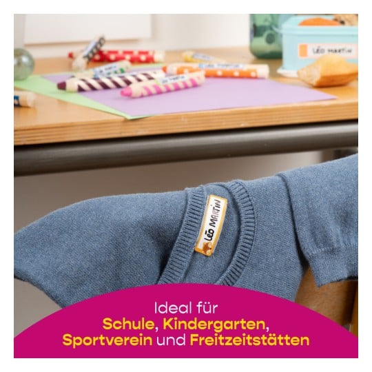 Das Bild zeigt einen blauen Pullover mit einem Etikett auf einem Tisch, der mit bunten Markern und Papieren in verschiedenen Farben überladen ist, was auf einen kreativen Raum für Kinder hindeutet.