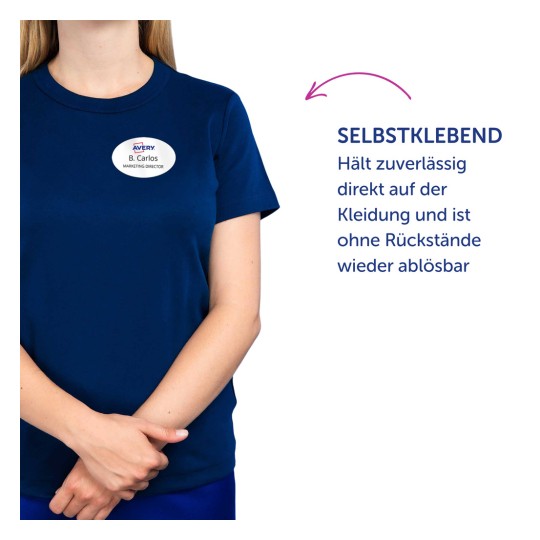 Das Bild zeigt eine Person, die ein marineblaues Hemd mit einem Namensschild trägt. Auf dem Namensschild steht 'Avery B. Carlos, Marketing Director'. Es gibt einen Text auf Deutsch, der besagt, dass das Namensschild selbstklebend ist, zuverlässig auf Kleidung hält und rückstandslos ablösbar ist.
