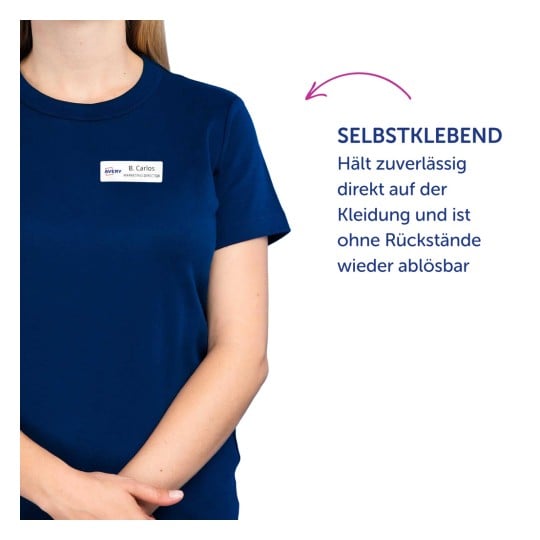 Das Bild zeigt eine Frau, die ein dunkelblaues T-Shirt trägt, mit einem Namensschild, auf dem 'B. Carlos Marketing Director' steht. Daneben gibt es einen deutschen Text, der darauf hinweist, dass das Etikett selbstklebend ist, zuverlässig auf Kleidung haftet und ohne Rückstände entfernt werden kann.