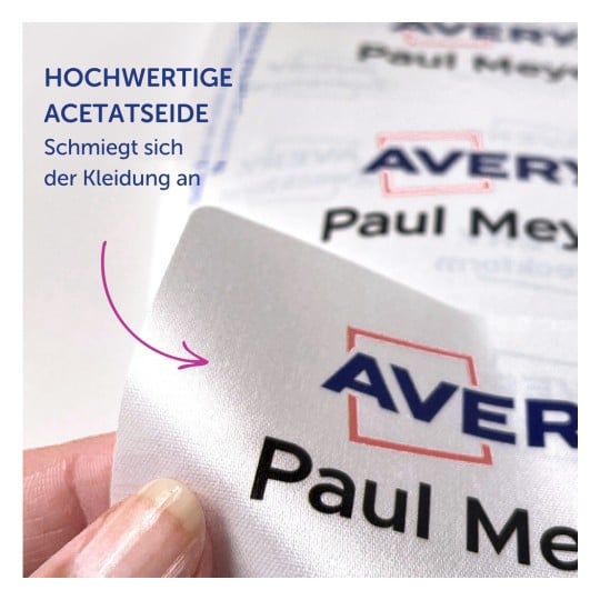 Das Bild zeigt eine Nahaufnahme eines Satinetiketts mit dem Text 'Avery Paul Meyer' und einer Beschreibung auf Deutsch über das Material, die darauf hindeutet, dass es gut auf Kleidung passt.