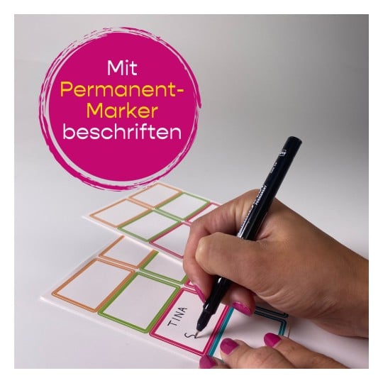 Das Bild zeigt eine Hand, die mit einem Permanentmarker auf ein Etikett schreibt. Das Etikett hat bunte Rahmen, und ein Text ist sichtbar, möglicherweise ein Name. Der Hintergrund ist schlicht und weiß.