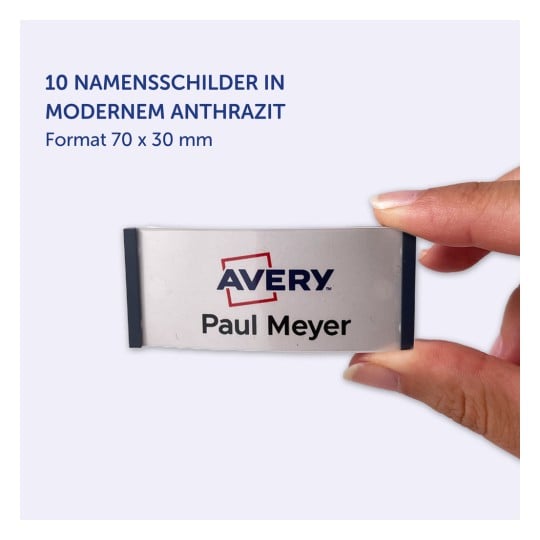 Das Bild zeigt eine Hand, die ein Namensschild mit dem Namen 'Paul Meyer' und dem Avery-Logo hält. Es zeigt an, dass es 10 Namensschilder in modernem Anthrazit gibt, Größe 70 x 30 mm.