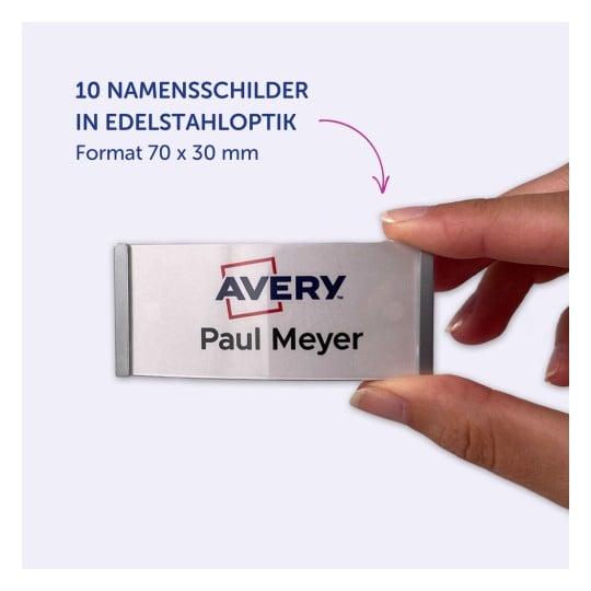 Das Bild zeigt eine Hand, die ein Namensschild von Avery hält. Das Schild trägt den Namen 'Paul Meyer' und ist in Edelstahloptik gestaltet, mit den Maßen 70 x 30 mm.