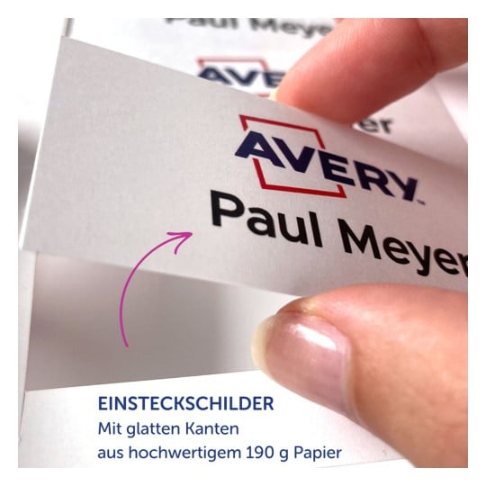 Das Bild zeigt eine Nahaufnahme einer Hand, die ein weißes Etikett mit dem Namen „Paul Meyer“ und dem prominent dargestellten „Avery“-Logo hält. Das Etikett scheint Teil einer Produktbeschreibung auf Deutsch zu sein, in der von glatten Kanten und hochwertigem Papier die Rede ist.