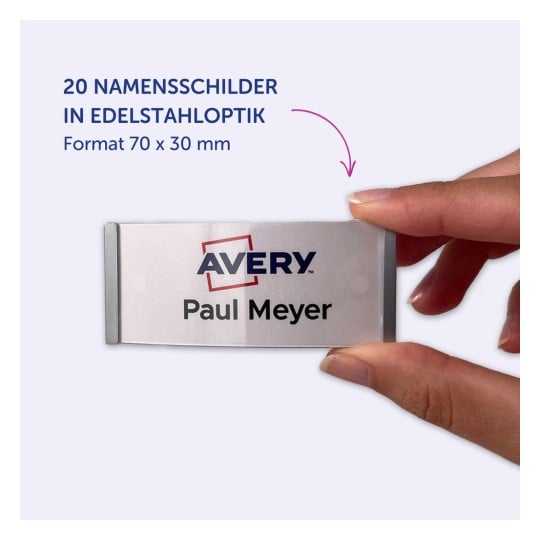 Das Bild zeigt eine Hand, die ein Namensschild hält, auf dem 'AVERY Paul Meyer' mit einer Edelstahloptik steht. Das Schild hat die Maße 70 x 30 mm und repräsentiert ein Set von 20 Namensschildern.