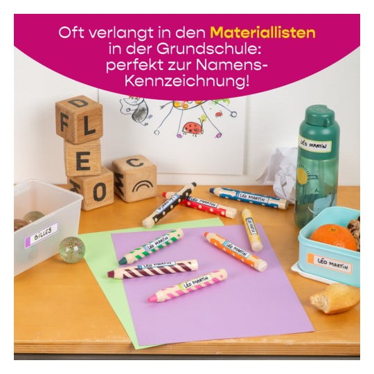 Das Bild zeigt eine schulszenische Szene mit bunten Buntstiften, die mit Namen beschriftet sind, einer Wasserflasche, einem Behälter mit Snacks und dekorativen Gegenständen wie Murmeln und Holzklötzen. Auf einem Tisch liegen Papiere in verschiedenen Farben ausgebreitet und an der Wand sind Zeichnungen zu sehen.