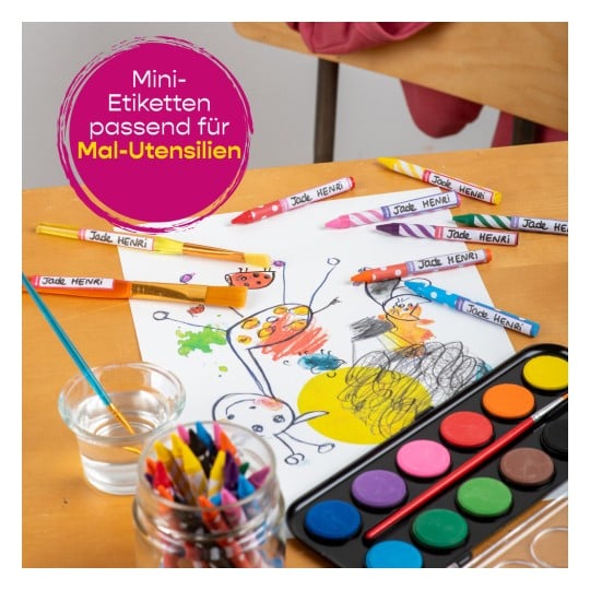 Das Bild zeigt ein Kunst-Setup mit Buntstiften, Wachsmalstiften, einem Zeichnungsblatt mit farbenfrohen Illustrationen, einem Wasserglas und einer Palette mit Aquarellfarben. Auf den Kunstmaterialien befinden sich kleine Etiketten mit der Aufschrift 'Jade HENRI'.