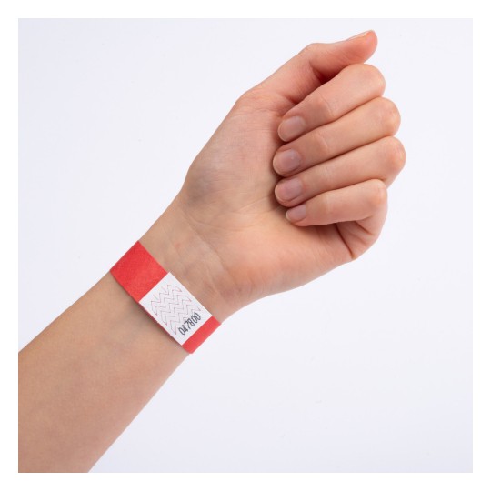 Das Bild zeigt eine Hand, die ein rotes Armband mit einem Zickzackmuster und einem kleinen aufgedruckten Etikett trägt, was darauf hindeutet, dass es möglicherweise zu Identifikationszwecken verwendet wird.