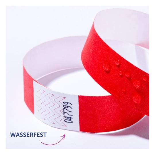 Das Bild zeigt ein rotes Armband mit einem weißen Abschnitt, der einige Markierungen aufweist, was darauf hinweist, dass es wasserdicht ist. Auf dem Armband befinden sich Wassertropfen.