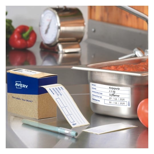 Das Bild zeigt eine Küchenszene mit einem Behälter Bolognese-Soße, einer Schachtel Avery-Etiketten, einem Stift und frischem Gemüse wie einer roten Paprika und einer Tomate.