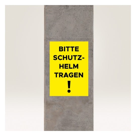 Das Bild zeigt ein gelbes Schild an einem Betonpfeiler mit der Aufschrift „BITTE SCHUTZ-HELM TRAGEN!“, was „Bitte einen Helm tragen!“ bedeutet und auf eine Sicherheitsanforderung hinweist.