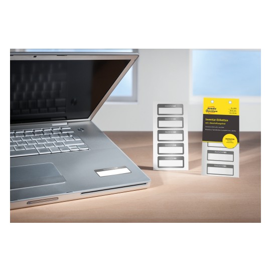 Das Bild zeigt einen Laptop mit einer reflektierenden Oberfläche und eine Packung mit der Aufschrift 'Inventar-Etiketten'. Die Etiketten sind weiß mit einem grauen Rand und sind für Bestandszwecke entworfen.