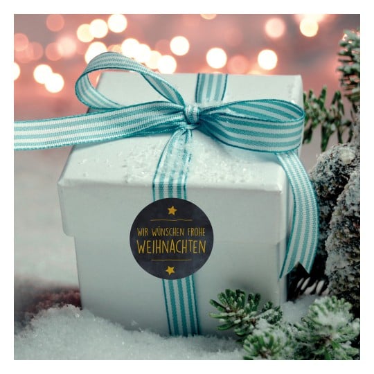 Das Bild zeigt eine weiße Geschenkbox, die mit einem blau-weiß gestreiften Band gebunden ist und auf einer schneebedeckten Oberfläche liegt. Sie hat einen runden Anhänger, auf dem 'Wir wünschen frohe Weihnachten' steht. Der Hintergrund ist weich verschwommen mit warmen, leuchtenden Lichtern.