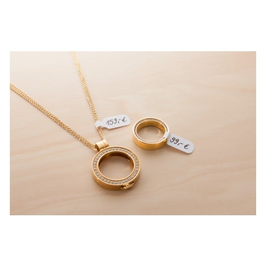 Das Bild zeigt eine goldene Halskette und einen goldenen Ring, die auf einer Holzoberfläche ausgestellt sind. Die Halskette hat einen runden Anhänger, der mit funkelnden Steinen verziert ist, und beide Artikel haben Preisschilder: 159 € für die Halskette und 99 € für den Ring.
