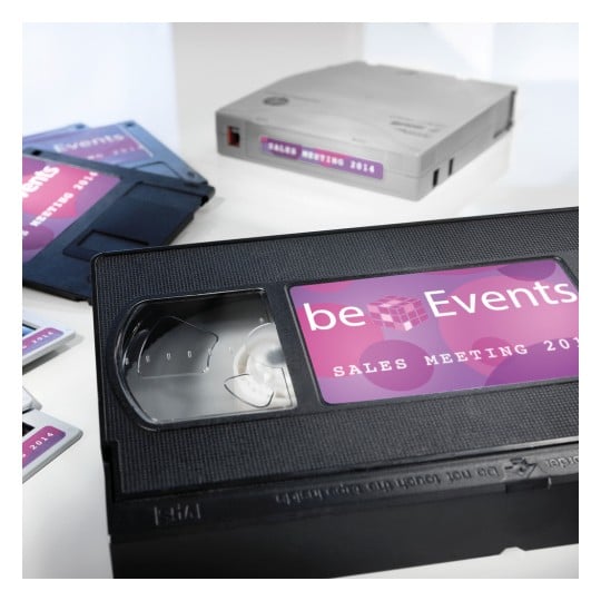 The image features a VHS tape labeled 'beEvents Sales Meeting 2014,' alongside several other media formats, including what appears to be a disk and another VHS tape, all arranged on a white background.
