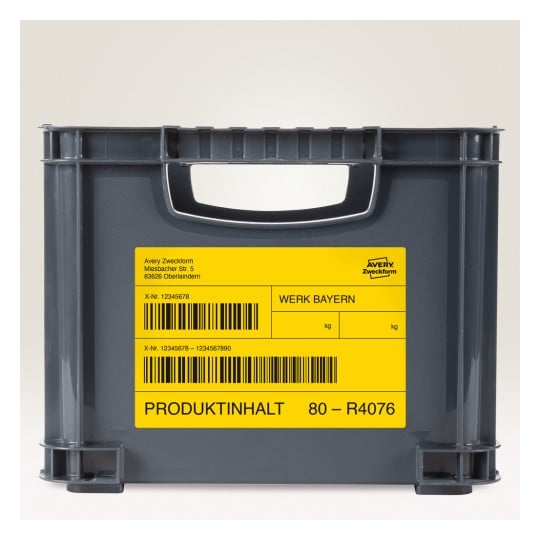 Das Bild zeigt eine graue Lagerkiste, die mit gelben Etiketten versehen ist. Die Etiketten enthalten Informationen wie 'Werk Bayern', einen Barcode und Produktdetails. Die Kiste scheint für Organisations- oder Versandzwecke verwendet zu werden.