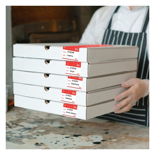 Eine Person in einer Kochuniform hält gestapelte Pizzakartons, die mit verschiedenen Namen und Bestellnummern beschriftet sind.