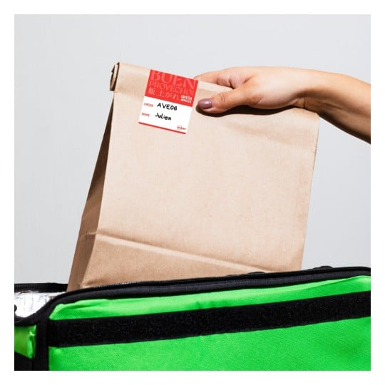 Eine Person hält eine braune Papiertüte mit einem Etikett darauf, das über einer grünen isolierten Liefer- bzw. Thermotasche platziert ist. Das Etikett enthält Details zur Bestellung.