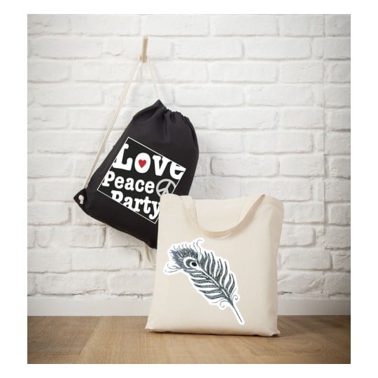 Das Bild zeigt zwei Taschen vor einer weißen Backsteinwand. Ein schwarzer Turnbeutel mit den Worten 'Love, Peace, Party' hängt dort. Daneben zeigt eine beige Tragetasche ein dekoratives Federdesign.